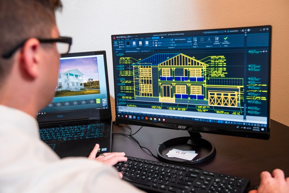 Ingénieur devant deux écrans, un modèle de maison et son plan technique ouverts dans un logiciel de dessin