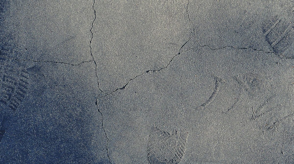 Dallage béton fissuré vu de dessus, réseau de fissures fines et traces de pas dans la poussière