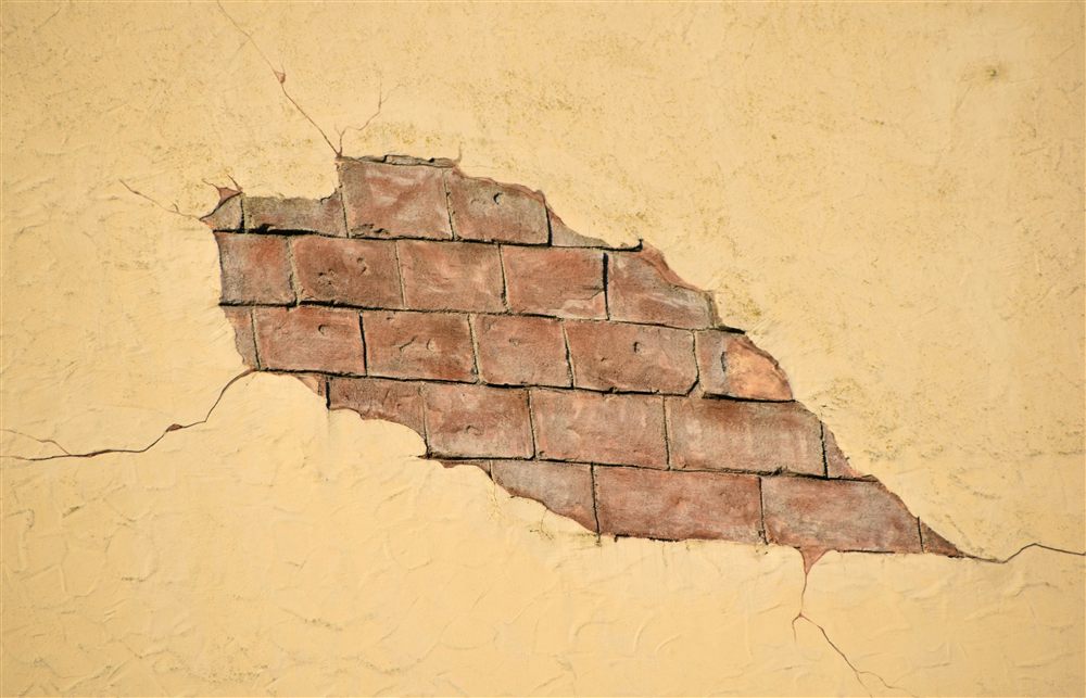 Enduit décollé et fissures sur un mur en maçonnerie, laissant apparaître les briques et les joints